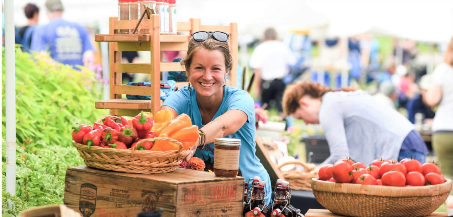 Woman in a light blue shirt with sunglasses on her head selling vegetables at a farm stand