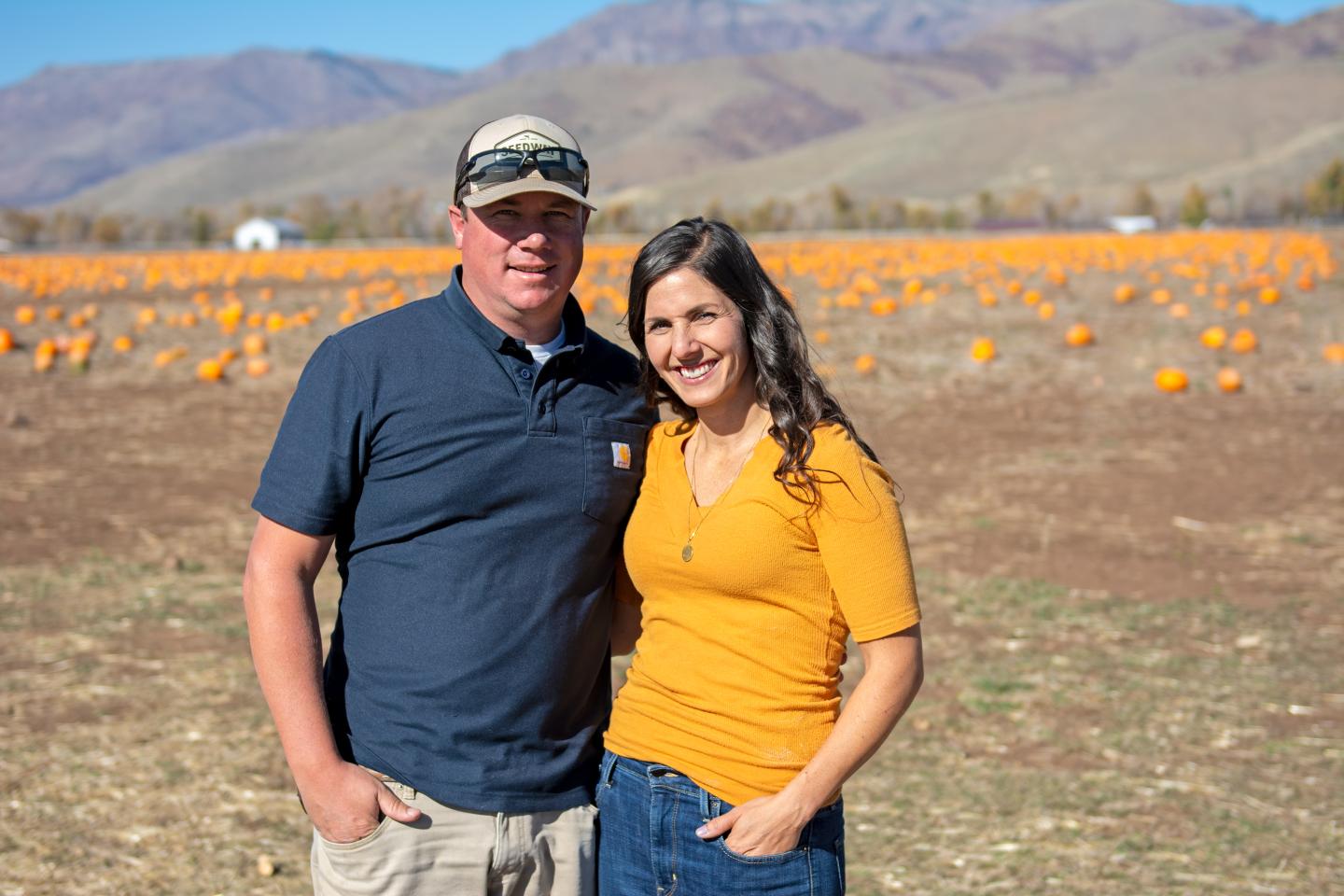 Two people smile at camera in front of rows of pumpkins and mountains