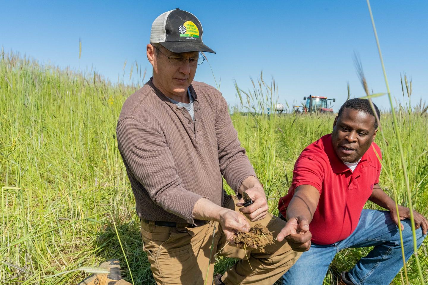 Two individuals kneeling in a field of grass. One has a handful of soil while the other points out soil health properties.