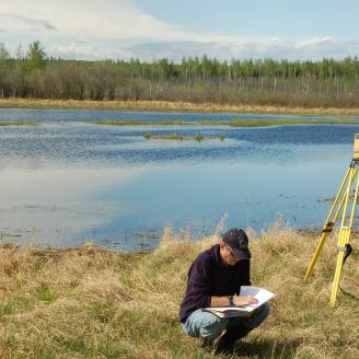 A man kneels down to write notes on a piece of paper next to survey equipment on a wetland in Alaska.
