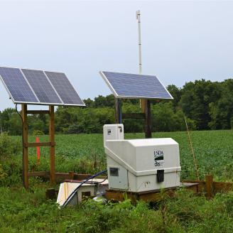 An edge-of-field research station for water quality testing in the foreground, with a cropland field in the background and gray skies above.
