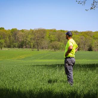 Man in a bright yellow shirt and grey pants standing in a field looking out at the farm property.