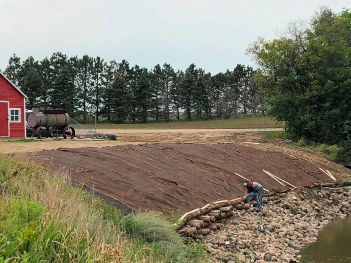 Technicians secure the turf reinforcement mat to the logs that had been previously placed in the riverbank to stabilize it. Rock riprap was laid below the logs to prevent the current from eroding the bank.
