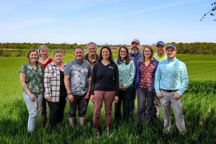 Group of men and women posing for a photo in a farm field on a sunny blue sky day.