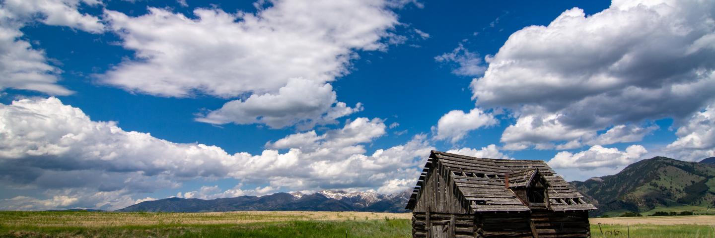 Photo of a log cabin in a large, grassy field. There are mountains several miles in the background some with snow on top, but most a green with grass and trees. The sky is partly cloudy
