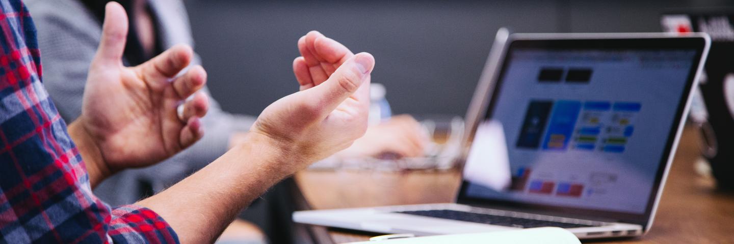 Caucasian hands in front of laptop, woman sits in background at a meeting