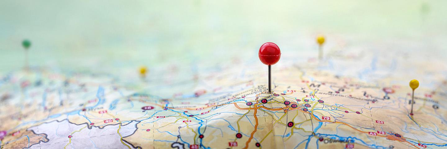 Red-headed pin stuck in roadmap; two yellow-headed and a green-headed pins in the background, stuck in other parts of the map