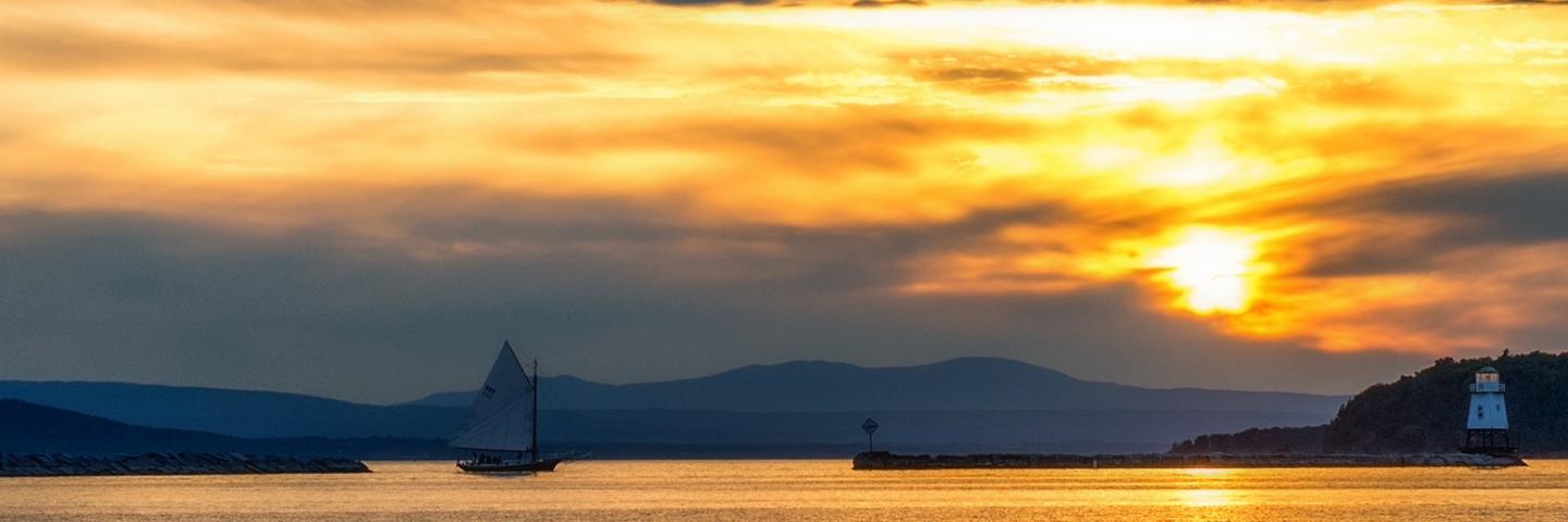 Small sailboat in water and lighthouse on island in Lake Champlain at sunset