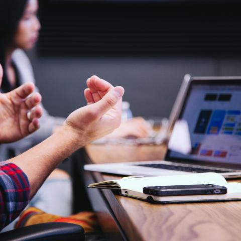 Caucasian hands in front of laptop, woman sits in background at a meeting