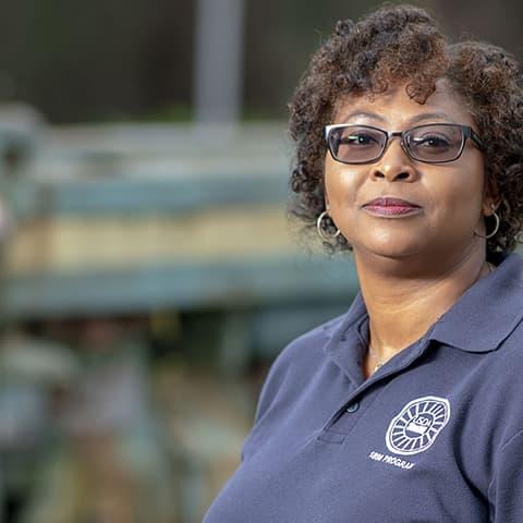 Photo of an African-American woman wearing glasses and a USDA 1890 Program polo shirt, with an old-fashioned John Deere tractor, grass, and a treeline in the background.