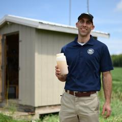 Dr. Chad Penn, Soil Scientist with USDA's Agricultural Research Service, stands facing the camera with a water sample in his hand. A related building is in the background, with a cropland field behind it and blue skies above.