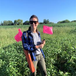 Person in a blue plaid shirt and sunglasses standing in a green field holding a pink flag marker.