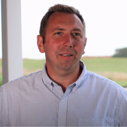White middle aged male wearing a blue and white vertical striped dress shirt in front ofa cow pasture