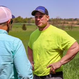 Man in a blue ballcap and bright yellow shirt talking to another man with a white ballcap and blue shirt.