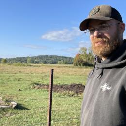 Man with a hat and beard standing in front of a pasture