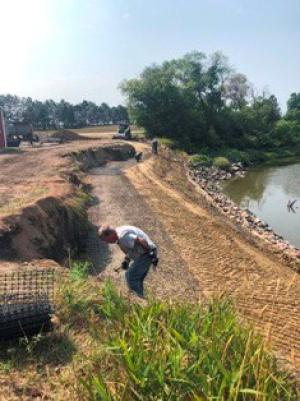 A construction team installs a geogrid stabilizer on the riverbank before the excavated area is backfilled with soil