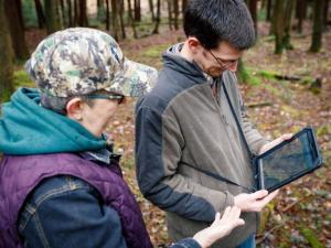 Two individuals standing in the woods, viewing information on a tablet.