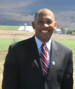 Dr. Terron Hillsman, NRCS SSRA Deputy Chief, stands smiling at the camera in a suit and tie with farmland in the background.