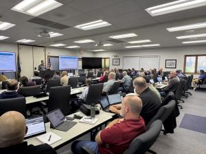 Full meeting with attendees focused on the speaker at the front of the room. 