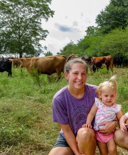 A family of two adults and two children squatted in their pasture with cows behind them.